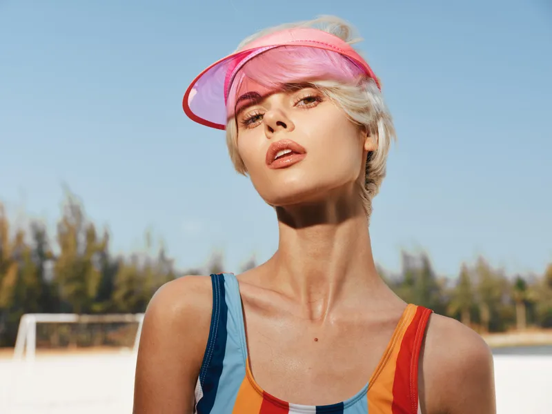 Close up portrait of a confident female model under bright sun, wearing a pink visor and colorful striped swimsuit top, against a clear blue sky and softly blurred athletic field.