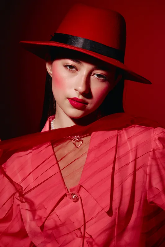 Young woman in stylish red hat and fashion dress looks confidently into the camera with soft dramatic lighting. Makeup is bold featuring red lips and flushed cheeks.