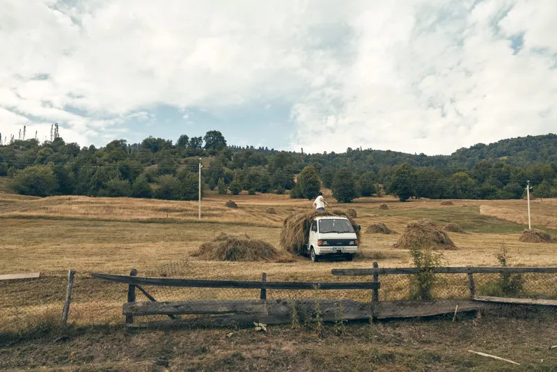 A white van rests in a wide open field beneath a cloudy sky, a weathered wooden fence in the foreground, creating a tranquil rural landscape with travel mood