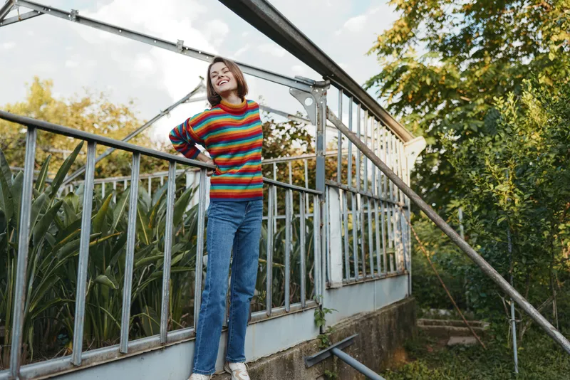 Woman in a colorful striped sweater and jeans, smiling and leaning on a greenhouse railing in an outdoor garden. Young adult with short bob, casual outfit, relaxed lifestyle fashion pose.