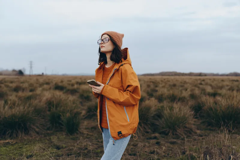 Woman lifestyle outdoor field grass autumn hat backpack walking countryside, young woman in orange jacket and knit beanie stands in a meadow, hands near chest, looking ahead, calm travel concept in