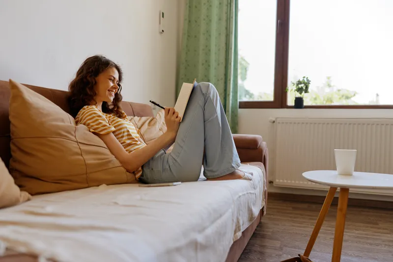 A young woman sits on a cozy sofa at home, reading a notebook with a pen in hand, smiling softly as warm daylight pours through a window, creating a relaxed mood.