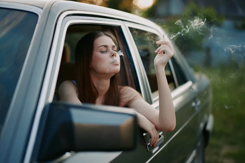A young woman with long brown hair is smoking while sitting in a vintage car, conveying a sense of relaxation and contemplation in a natural setting.