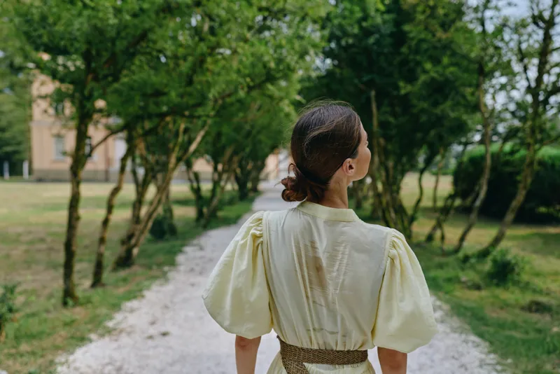 Woman back walking on park path among trees in dress, authentic inclusive portrait of nature stroll with calm mood