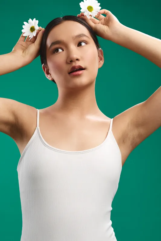 asian woman wearing a white tank top poses in a studio setting with daisies in her hair, presenting a bright, calm vibe against a vivid green backdrop.