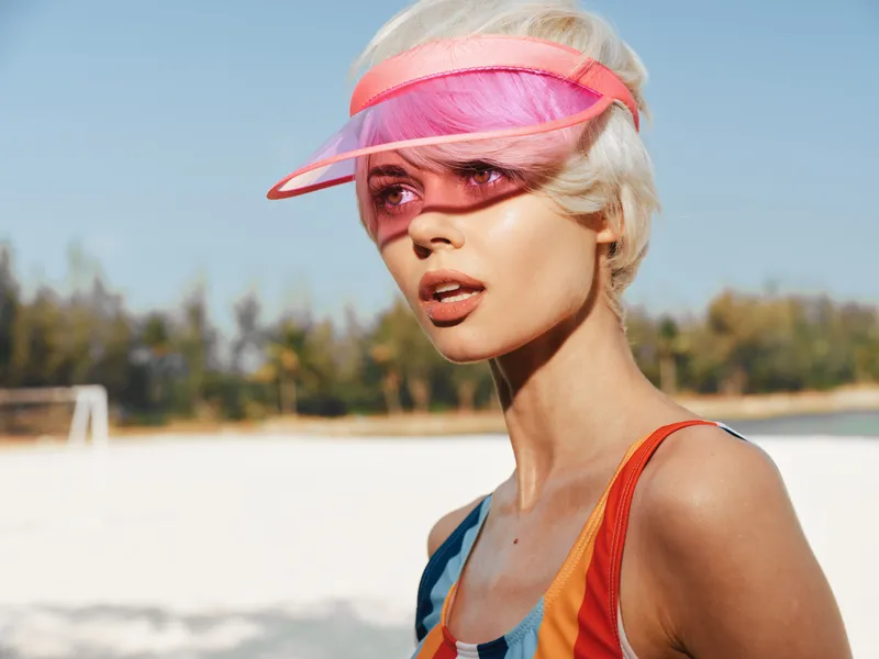 Portrait of a stylish woman at a sunny beach wearing a colorful swimsuit and a pink visor, showcasing bold summer fashion with soft wind, bright light, and a confident, carefree attitude.