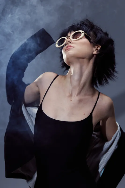 Stylish woman wearing sunglasses and a black dress posing confidently with smoke on dark background.