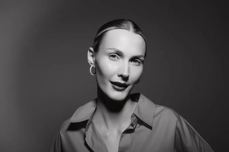 Portrait of a young white woman with fair skin and slicked back hair wearing a collared shirt, smiling softly in black and white studio lighting.