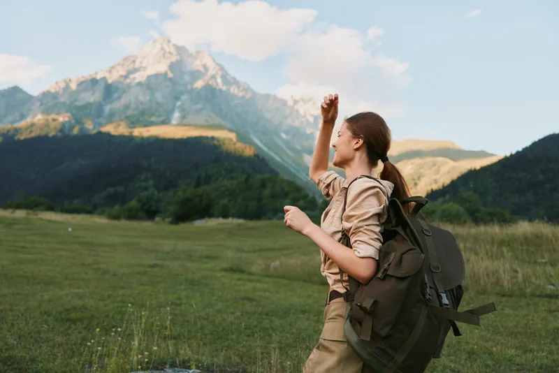 Cheerful woman hiking outdoors, raising a fist in celebration, wearing a backpack, in a lush valley with distant mountains, bright sky and natural colors