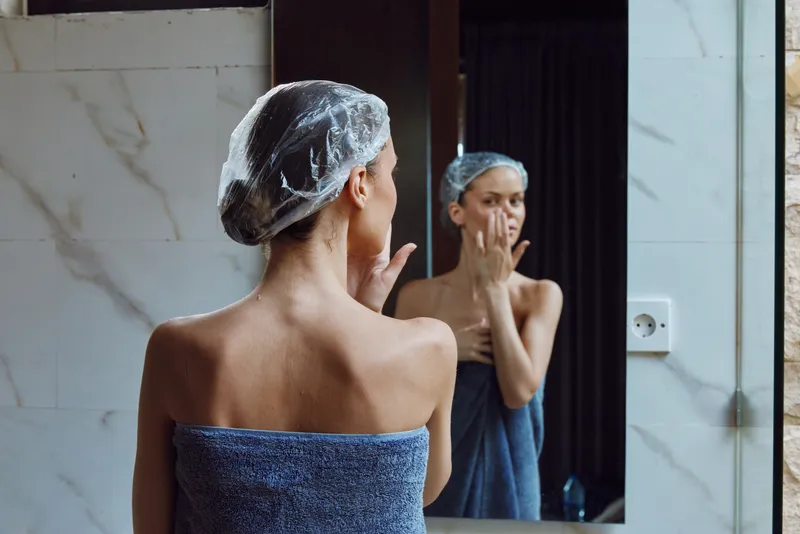 woman in towel applying skincare cream on face in bathroom, wearing shower cap, natural light, morning routine, self-care