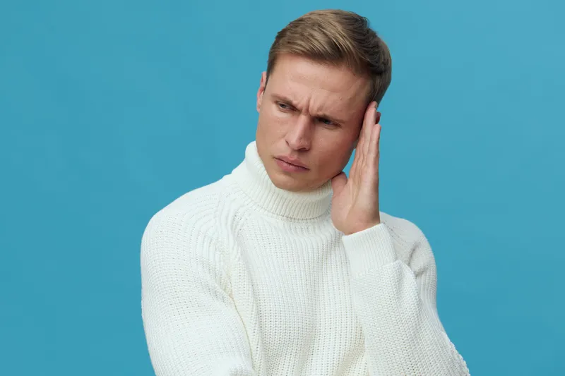 Thoughtful young man with short hair wearing white sweater isolated on blue background studio portrait, contemplating or listening carefully, casual style, emotion of curiosity and concern, appealing 