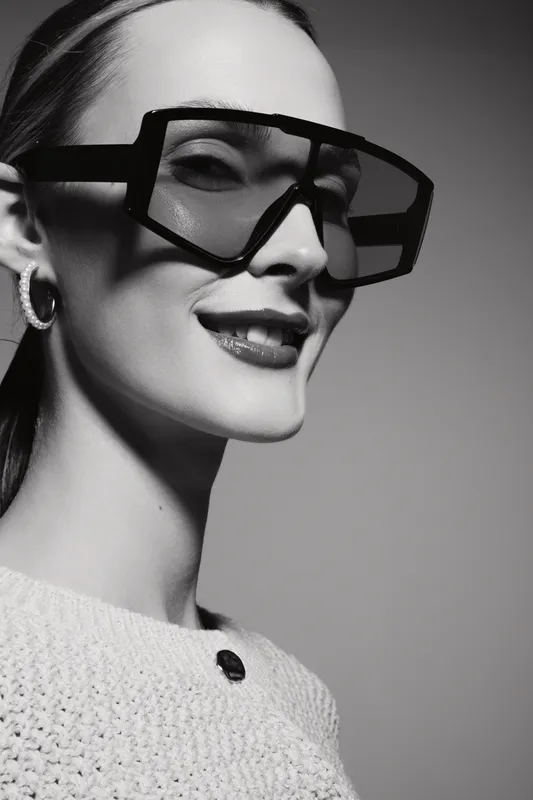 Portrait of a fair skinned Caucasian woman wearing large geometric sunglasses and a textured sweater, smiling confidently against a simple studio background.