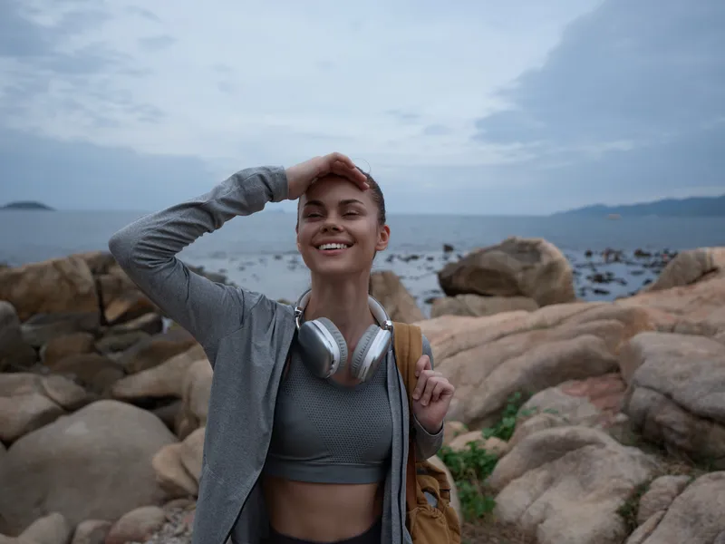 Young woman enjoying a fun outdoor activity on rocky beach, wearing sportswear and headphones, with a joyful smile and a serene ocean background