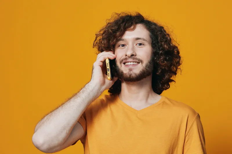 Young man with curly hair smiling while talking on the phone against a vibrant yellow background, wearing a casual orange t shirt that evokes a cheerful, warm feeling