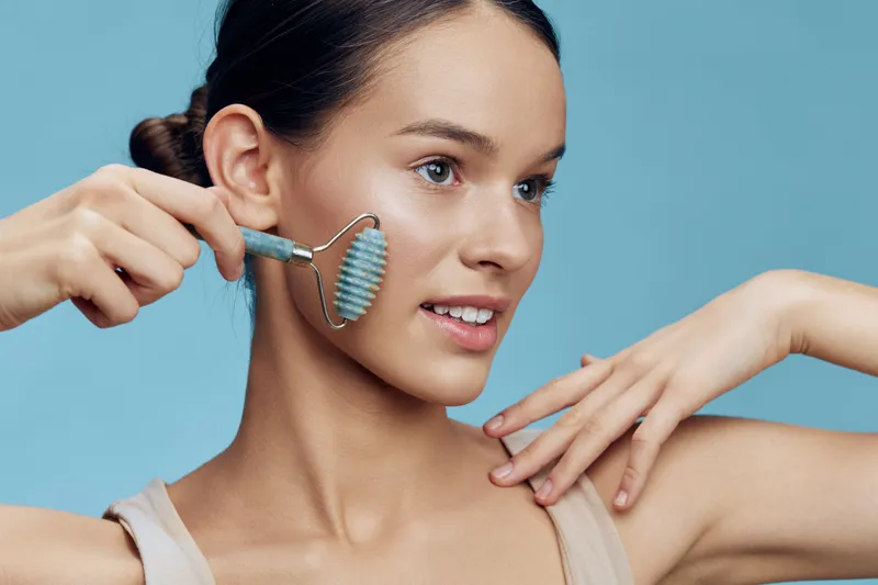 Young woman using jade roller on her face for skincare, showcasing smooth skin and glowing complexion against a soft blue background