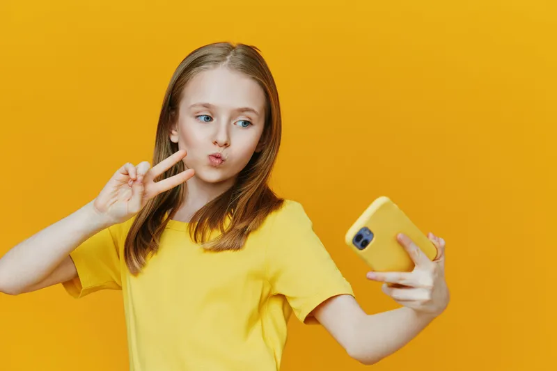 Cheerful girl taking a selfie on a bright yellow background, wearing a trendy yellow shirt, expressing fun emotions with a peace sign gesture, perfect for social media themes
