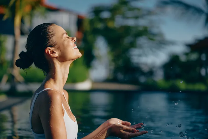 Woman outdoors, sun on her face, water and pool behind, relaxation and joy in a sunlit, candid moment, gentle breeze, natural colors, carefree mood