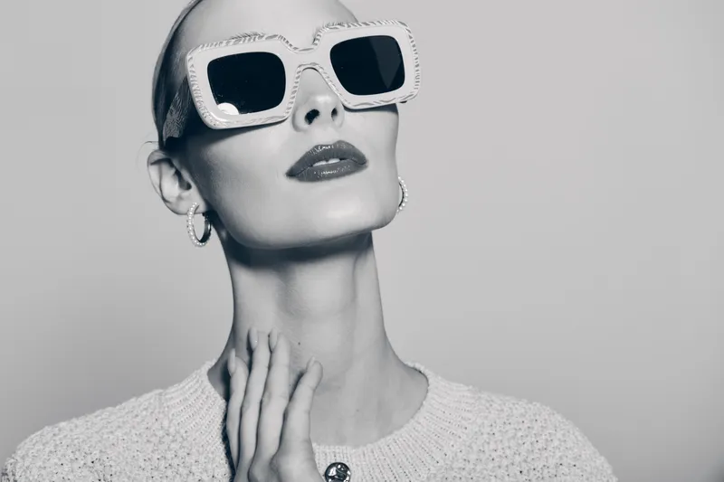 Stylish caucasian woman with pale skin wearing white sunglasses and knitted sweater poses with hands near neck in monochrome studio portrait.