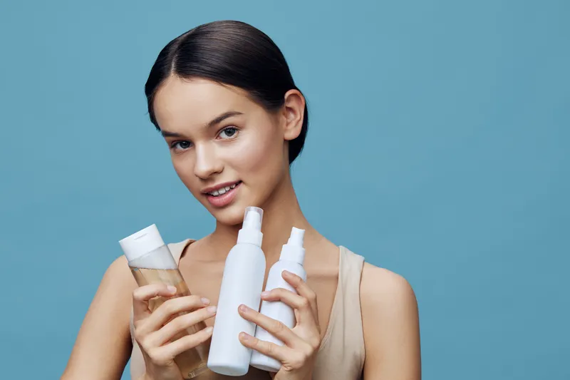Young woman with a natural look holding hair care products against a blue background, showcasing healthy hair and a skincare routine for vibrant confidence