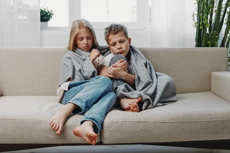 Two children wrapped in gray blankets sit closely on a beige sofa, looking unwell and tired in a bright living room. Concept of childhood illness and care at home.