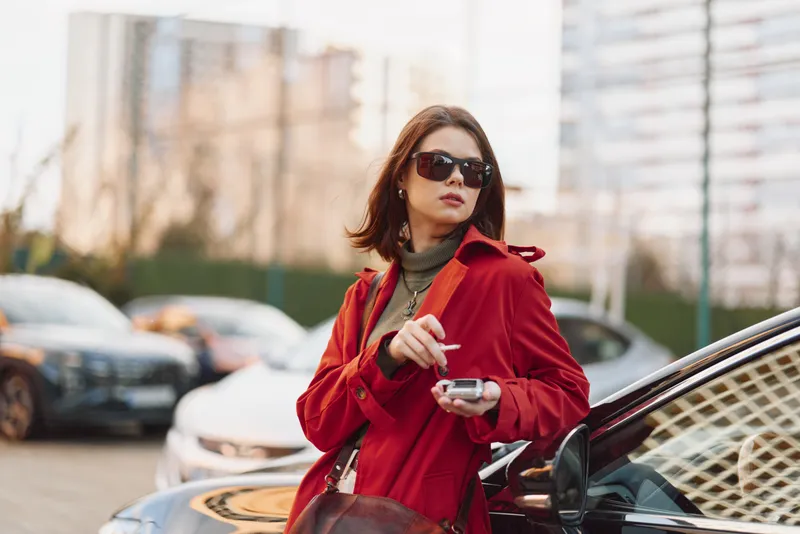 A young woman in a red coat and sunglasses stands by a car outdoors holding a smartphone while looking to the side with a confident expression.