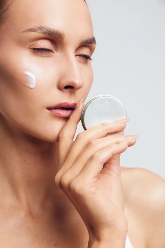 Young woman with closed eyes applies cream on her cheek holds cosmetic jar near face skincare beauty routine natural fresh skin isolated on white background closeup portrait