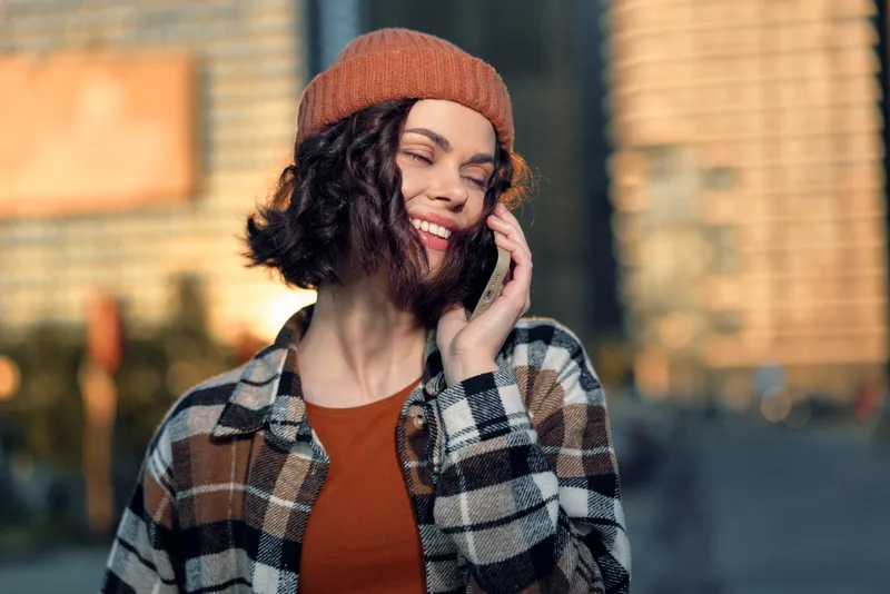 woman smile on phone in city at golden hour, beanie and plaid shirt, candid lifestyle portrait conveying authenticity, joyful mindful living and emotional storytelling in urban glow