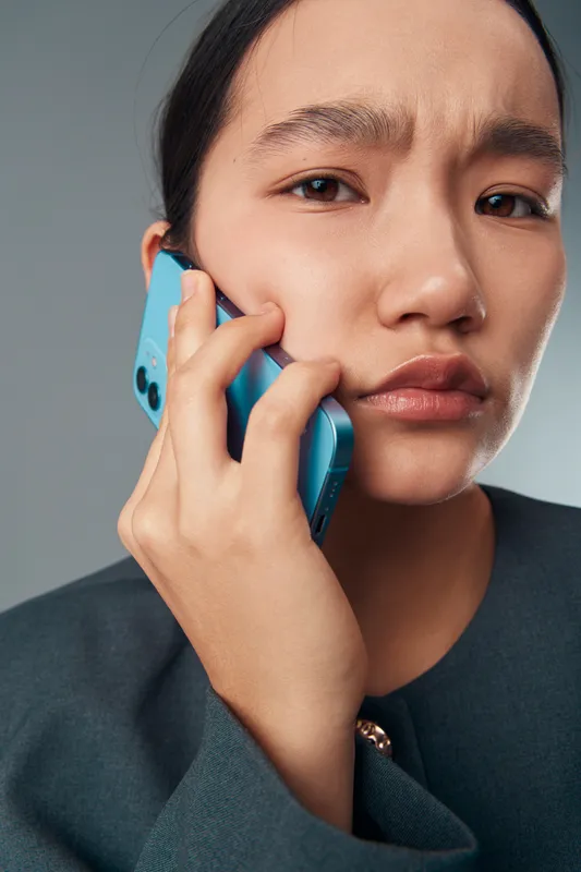 Young woman holding blue smartphone near ear with serious expression, closeup portrait on grey background, communication, technology, connection and mobile device usage concept.