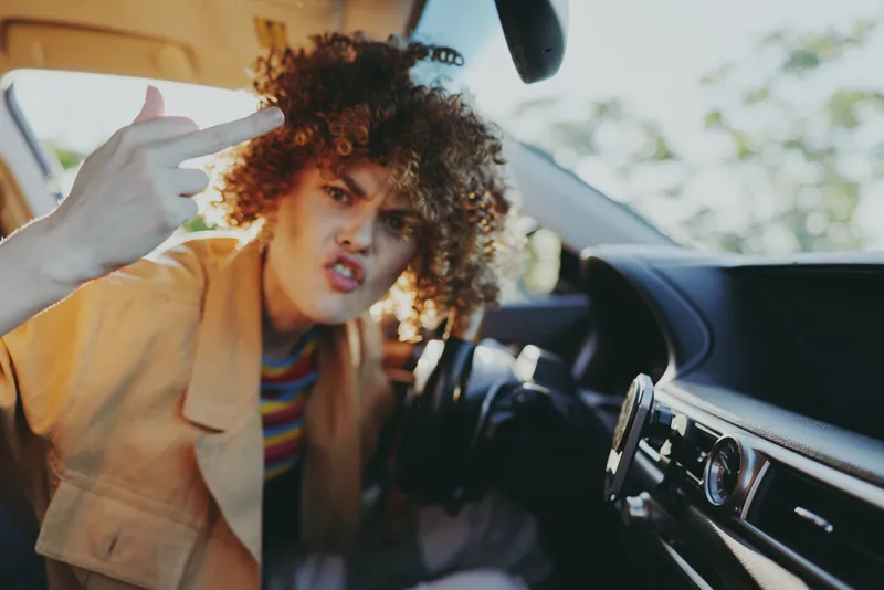 Woman in a car, retro film color, bold expression, middle finger gesture, casual jacket, curly hair, street vibe, candid lifestyle moment inside vehicle, vibrant street scene
