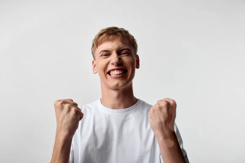 Smiling, excited young man raising clenched fists in celebration, conveying victory and confidence in a bright studio setting, dynamic mood for motivation and success campaigns