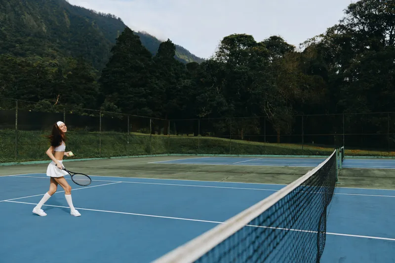 Young female tennis player enjoying a game on a blue court surrounded by lush greenery and mountains, expressing joy and athleticism in a summer setting