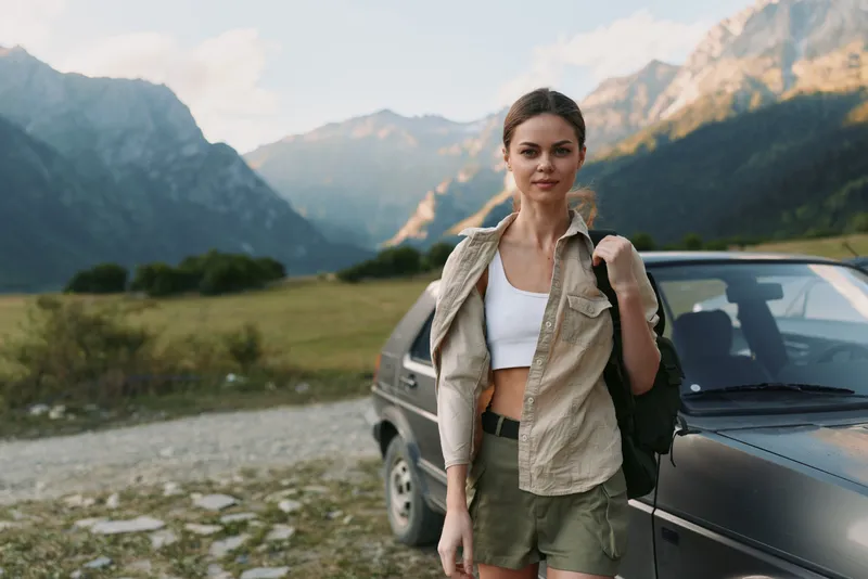 Young woman with a backpack stands beside a car on a mountain road enjoying the outdoor adventure and scenic landscape during a casual hike.