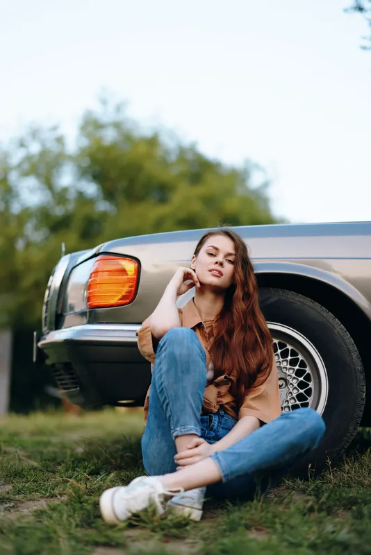 Young woman with long hair sitting by a vintage car, wearing casual clothing, expressing a thoughtful emotion, outdoor setting with greenery and a vintage vibe.