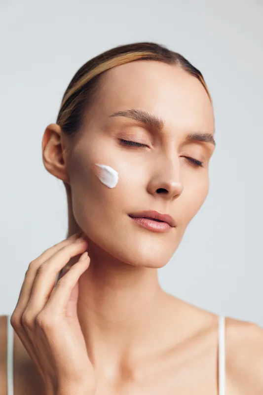 A young woman with closed eyes applies white cream on her cheek in a minimal studio setting. The image represents skincare, beauty routine, and healthy skin care concept.