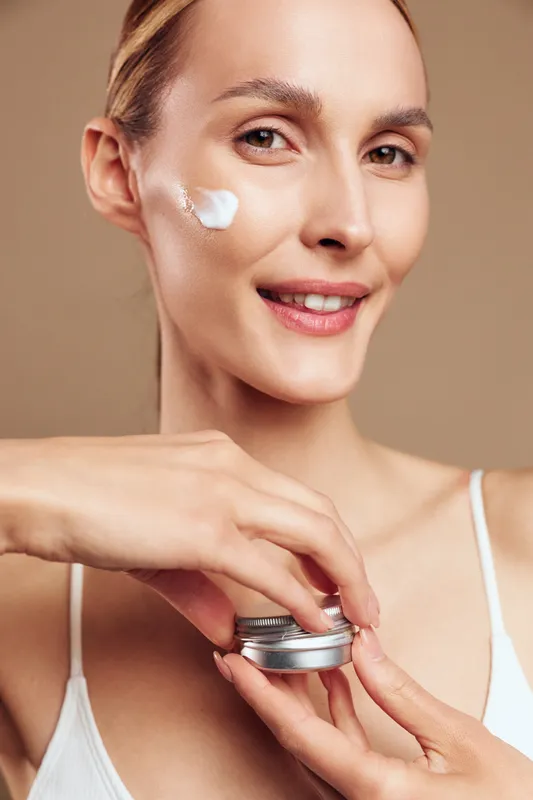 Young woman with clear skin applying white face cream on cheek, smiling gently. Concept of skincare, beauty routine, and natural wellness. Soft lighting highlights healthy skin texture.