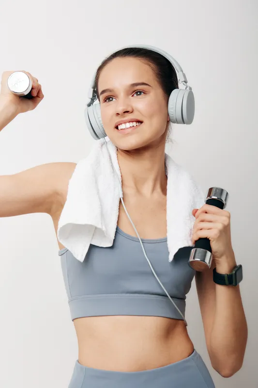 Fitness woman lifting dumbbells with headphones and a towel around her neck, mid workout in a bright studio, showcasing strength, focus, and an active lifestyle.