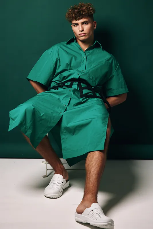 Young man with curly hair wearing green oversized shirt dress and white slip-on shoes sitting against dark green background in studio with casual modern fashion style.