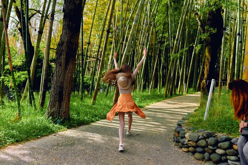 girl skirt hat bamboo path running along a gravel trail in a sunlit bamboo forest, carefree summer motion, back view of joyful young woman exploring nature outdoors