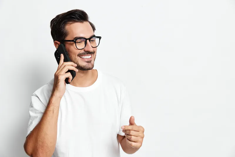 Smiling man wearing glasses holds smartphone while talking, showcasing a casual and cheerful vibe against a minimalistic solid background. People lifestyle concept.