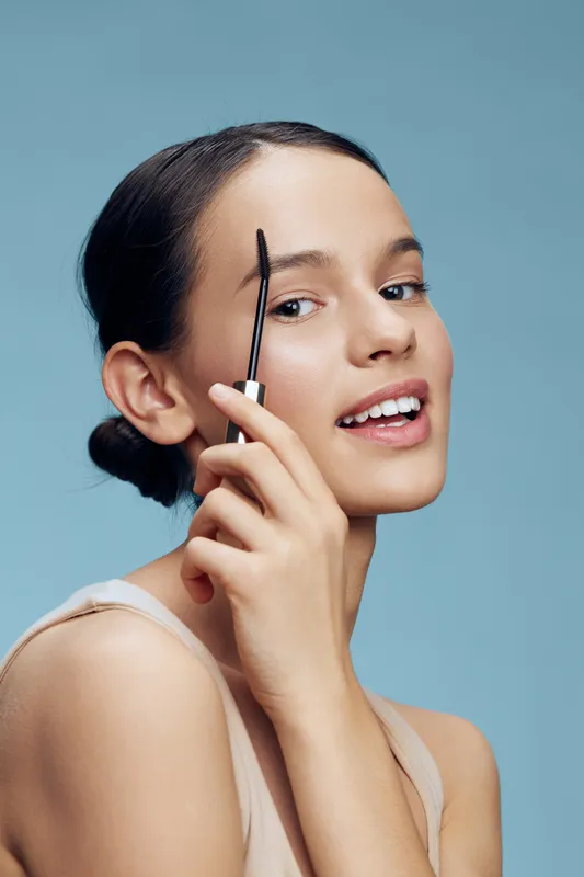 Young woman applying eyebrow makeup with a smile against a soft blue background, showcasing beauty and confidence in skincare routines