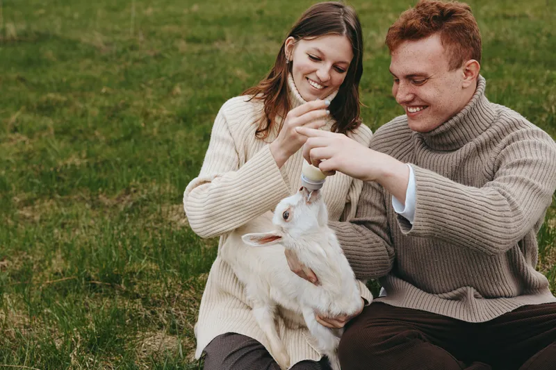 A loving couple outdoors feeds a young goat kid with a bottle, smiling softly as the animal drinks, in a grassy field, capturing warm countryside care and companionship