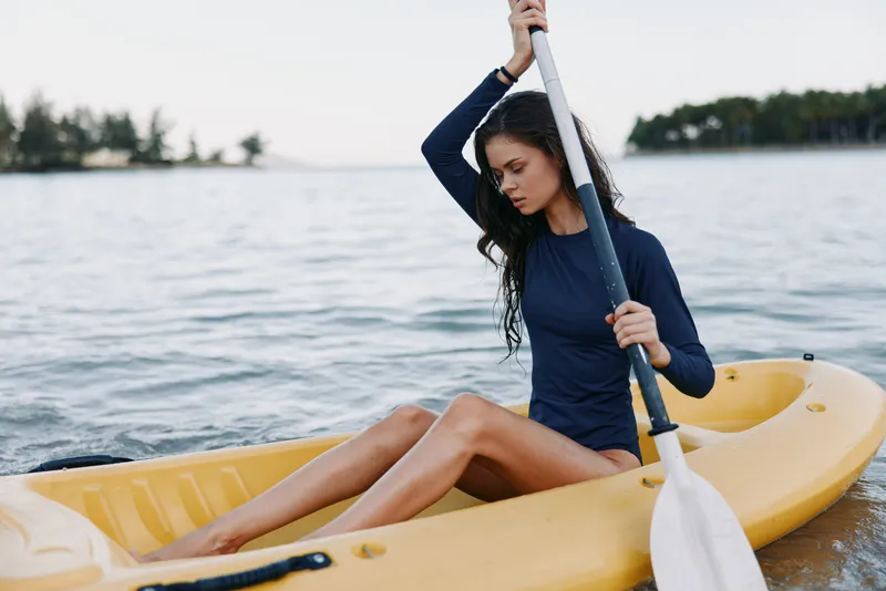 Young woman paddling yellow kayak on calm lake, wearing navy long-sleeve top, with focused expression and scenic forest background.