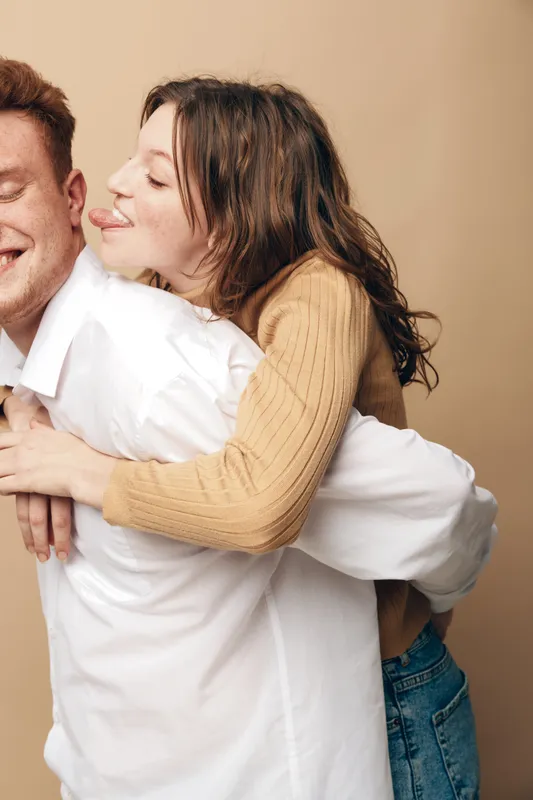 Happy young woman in beige sweater playfully hugs smiling man from behind, sticking out her tongue. Casual couple having fun together on neutral background. Concept of love and joy.