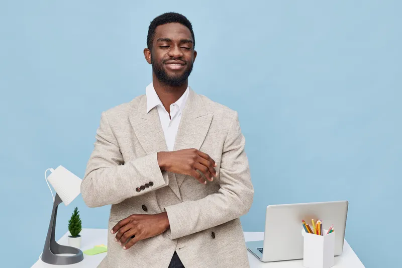 Confident young African man in beige blazer and white shirt smiling and looking at camera while standing at modern workspace with laptop and stationery on light blue background. Business success conce