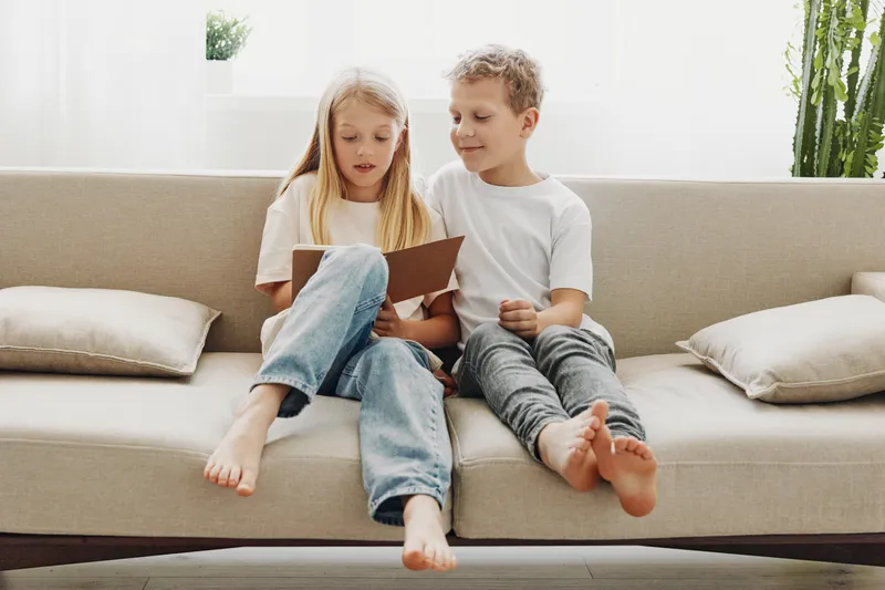 Two children sitting barefoot on a beige sofa reading a book together in a bright living room with natural light. Concept of childhood, education, and family bonding at home.