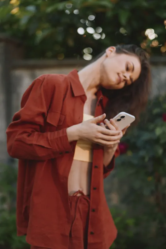 Woman with long hair wearing a rust red shirt using a smartphone outdoors in a garden. Casual fashion, lifestyle moment highlighting connectivity and relaxed mood and authenticity