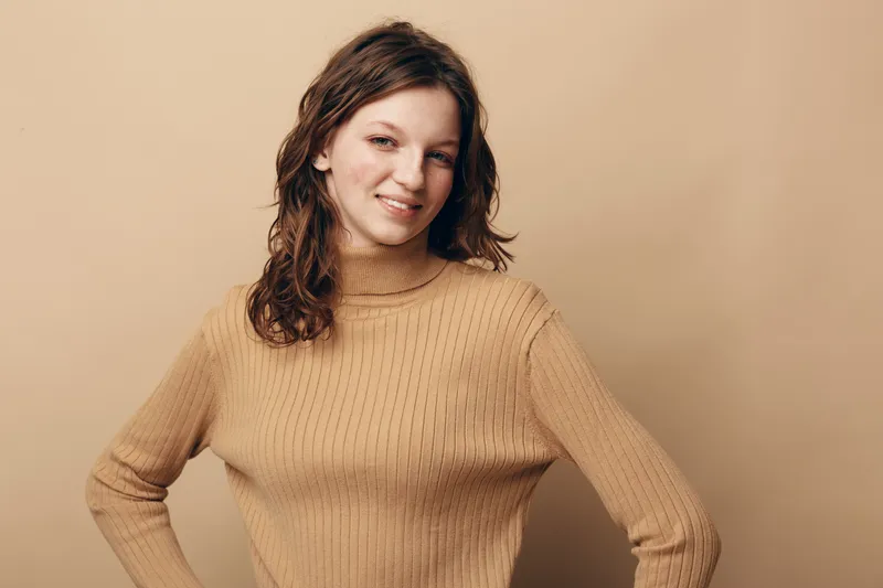 Young woman with wavy brown hair wearing beige knitted sweater smiles confidently with hands on hips against plain beige background. Concept of casual style and natural beauty.