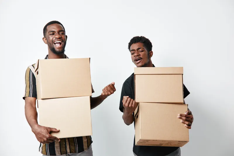 Two young black men with dark skin holding cardboard boxes and smiling on white background. African ethnicity, casual clothing, moving or delivery concept with happy expression.