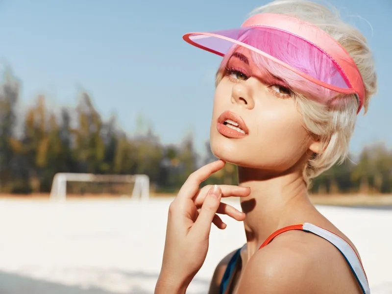 Bright portrait of a young woman wearing a pink visor under a clear sun, gazing softly at the camera, lips slightly parted, evoking a casual, sunny summer vibe.