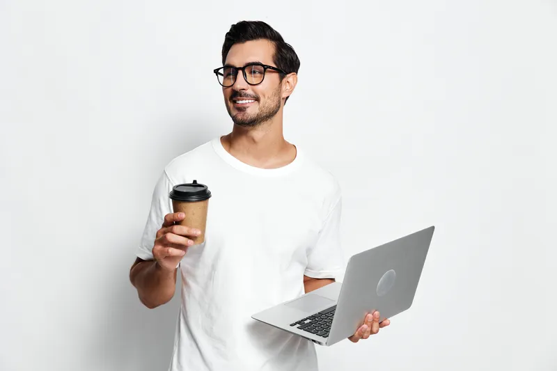 Smiling man holding a coffee cup and a laptop, wearing glasses, standing in front of a plain white background. Professional lifestyle concept.